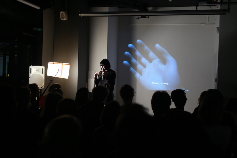 A photograph from an Interfaces Monthly event with a woman with short dark hair speaking into a microphone in front of a shadowy crowd with an illuminated blue hand projected onto the wall behind her.