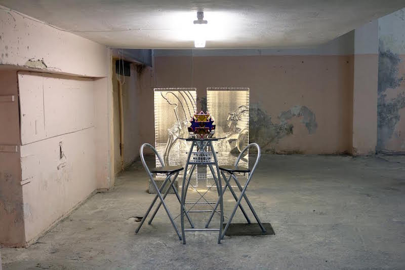 An Iain Ball image of a high glass table and two high chairs in an empty warehouse space. To the left there is peeling plaster on the wall and white plasterboard storage unit. There is an exposed light hanging from the ceiling above the table.