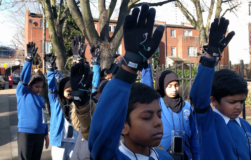 A Ling Tan photograph of 8 school children in uniform aged around 10 years old raise their right hands in the air with their eyes closed. On their rights hands they are wearing black gloves with wires coming out. In the background are three trees and a block of red brick buildings. 