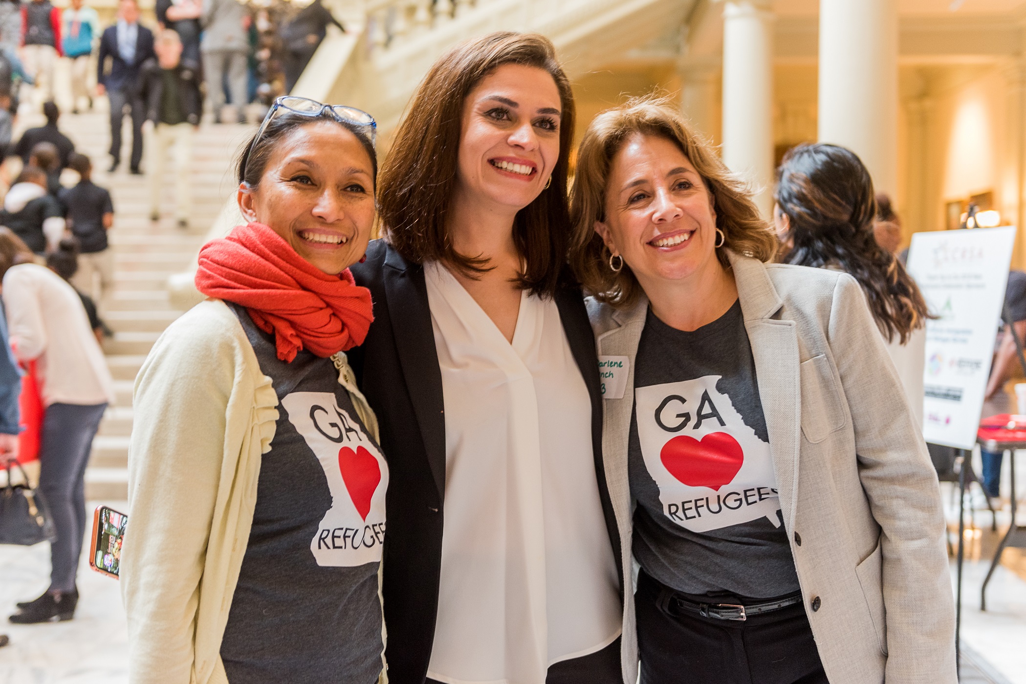 Three women wearing GA Loves Refugees t-shirts smile together at the Georgia State Capitol.