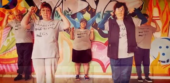 This is an image of part of Mambourin's hip hop crew. it features four people. All wearing grey tshirts with the words 'Mambourin Leaping Legs' in the middle of them written in black capital letters. There are two people standing in the front with their arms up and two people standing in the back with the arms up. Everyone has their fists clenched. They are dancing.