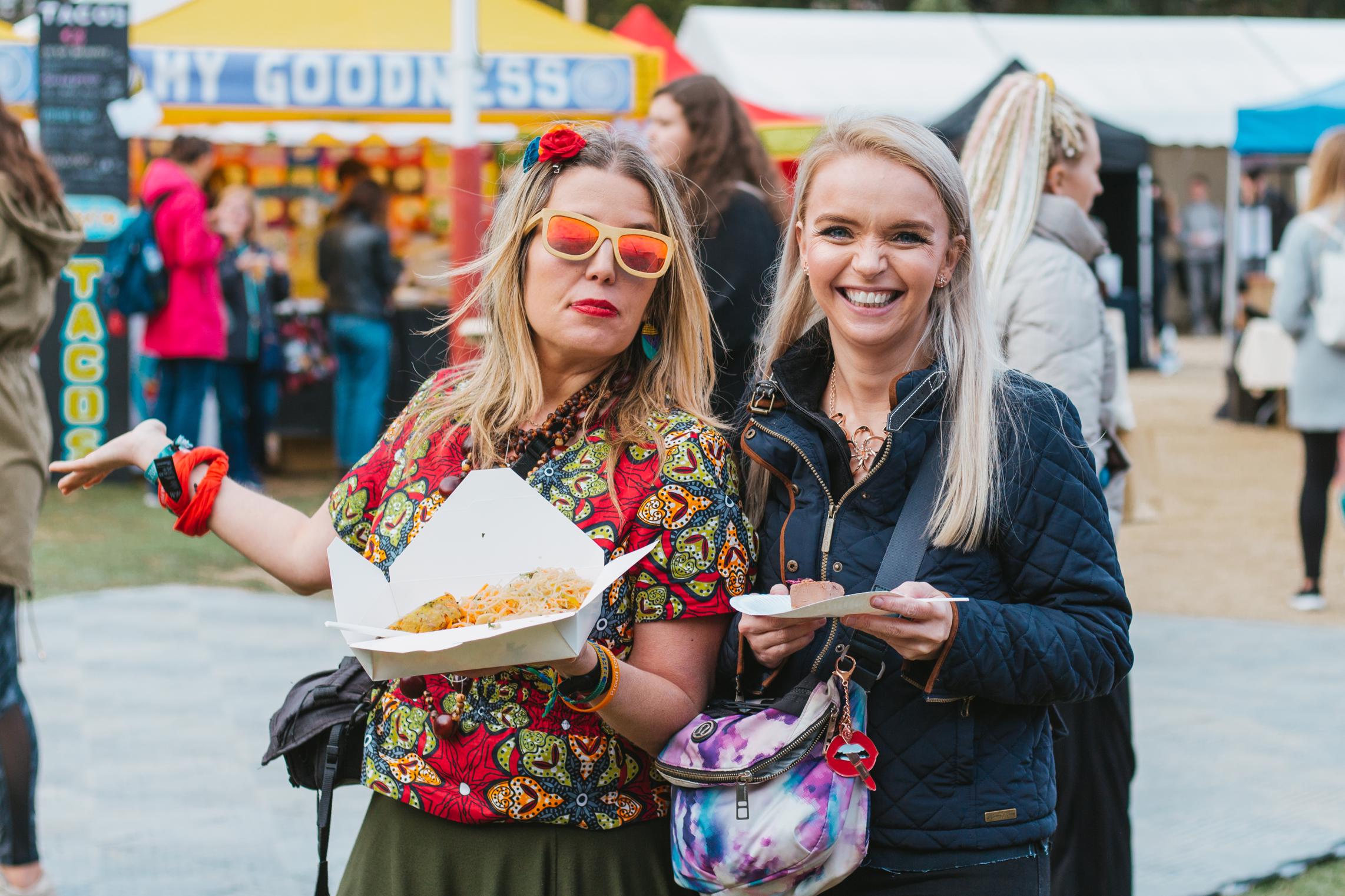 A WOMAN WEARING MIRROR SUNGLASSES INA COLOURFUL OUTFIT HOLDING VEGAN FOOD WITH ANOTHER WOMAN ALSO HOLDING FOOD SMILING AND LAUGHING AT DUBLIN VEGFEST