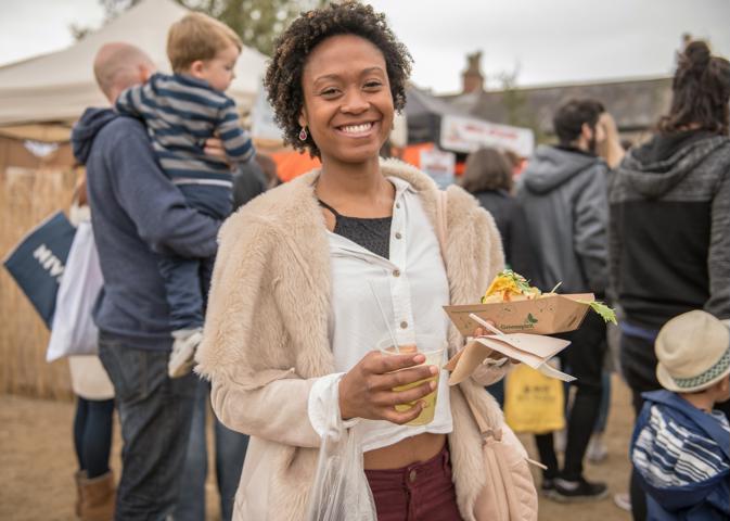 A black woman smiling, standing in front of some moving people carrying a child at shoulder level at Vegfest holding a paper boat of vegan food and a compostable glass full of sparking kefir drink.