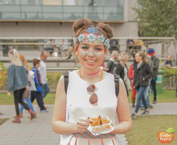 YOUNG WOMAN SMILES, WEARING WHITE FESTIVAL DRESS, TIARA OF BLUE FLOWERS AND FACE MAKE UP OF WHITE STARS AND SPARKLES STANDS OUTSIDE AT DUBLIN VEGFEST HOLDING A PLATTER OF VEGAN FOOD.