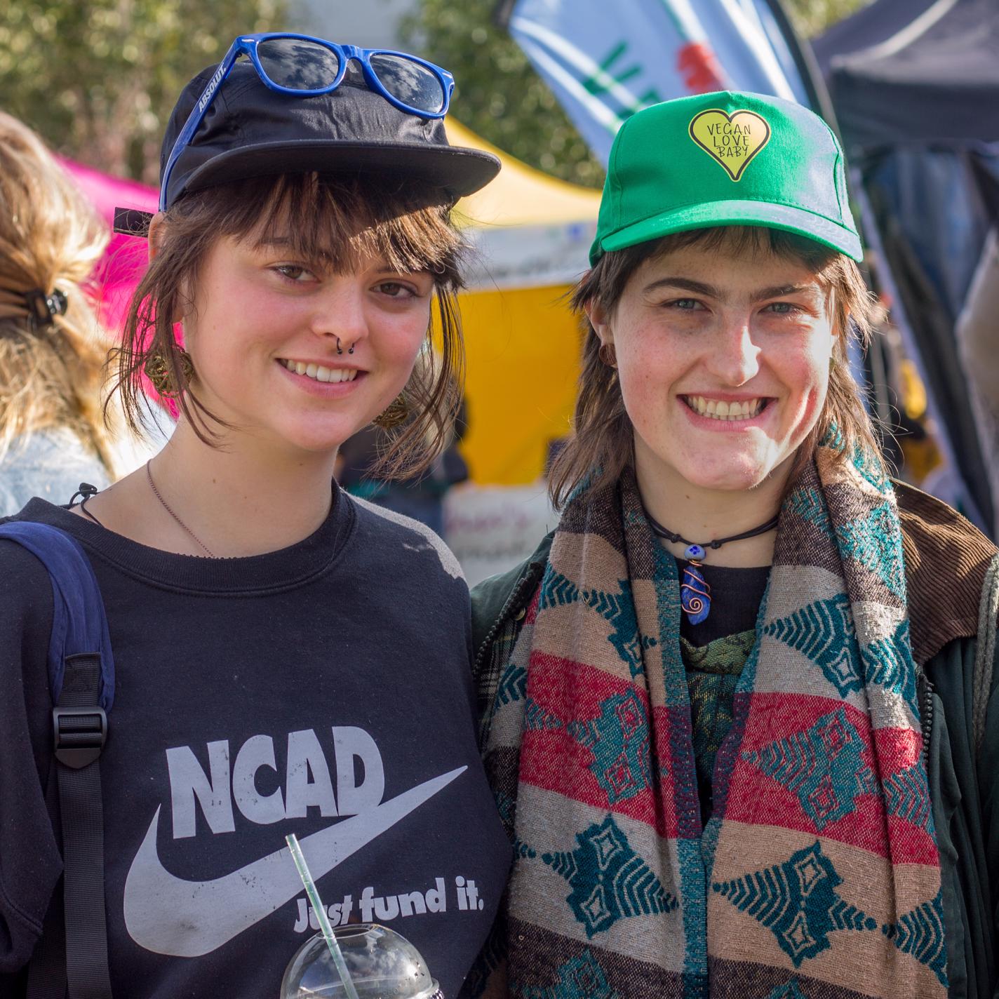 TWO YOUNG WOMEN SMILING. WEARING TRUCKER/BASEBALL CAPS