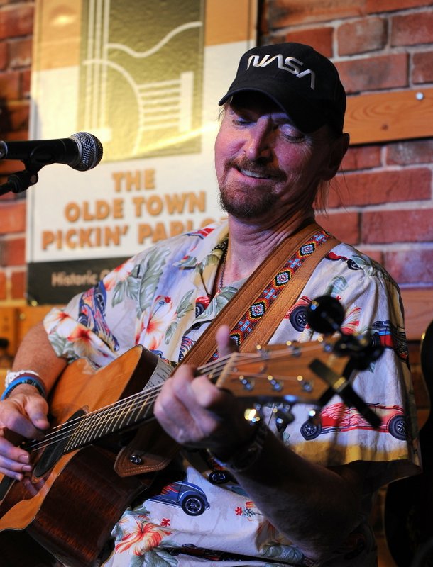 a goateed man with pulled back hair underneath a black NASA baseball cap looks loving at the acoustic guitar he is strumming. he is wearing a light buttoned up shirt with hawaiian print and flaming cars on it.