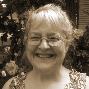 a white haired woman with bangs and glasses grins at the camera with a backdrop of leaves and what appears to be a house. she is wearing a floral ruched tank top