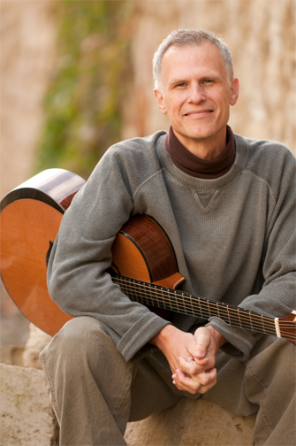 a gray haired man with a gray sweater sitting on a ledge, his hand clasped and a guitar nestled under his right armpit