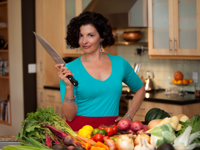 a woman with short brown hair with a blue shirt and her hand on her hip, hold a chopping knife before a layout of many fruits and veggies