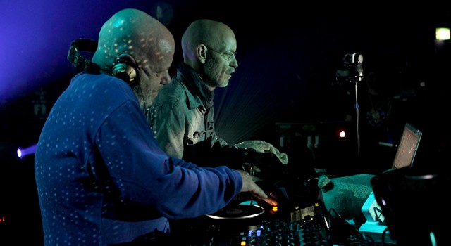 A view from behind two bald men hovering over computer screens and mixing boards on a stage before a huge group of dancing people