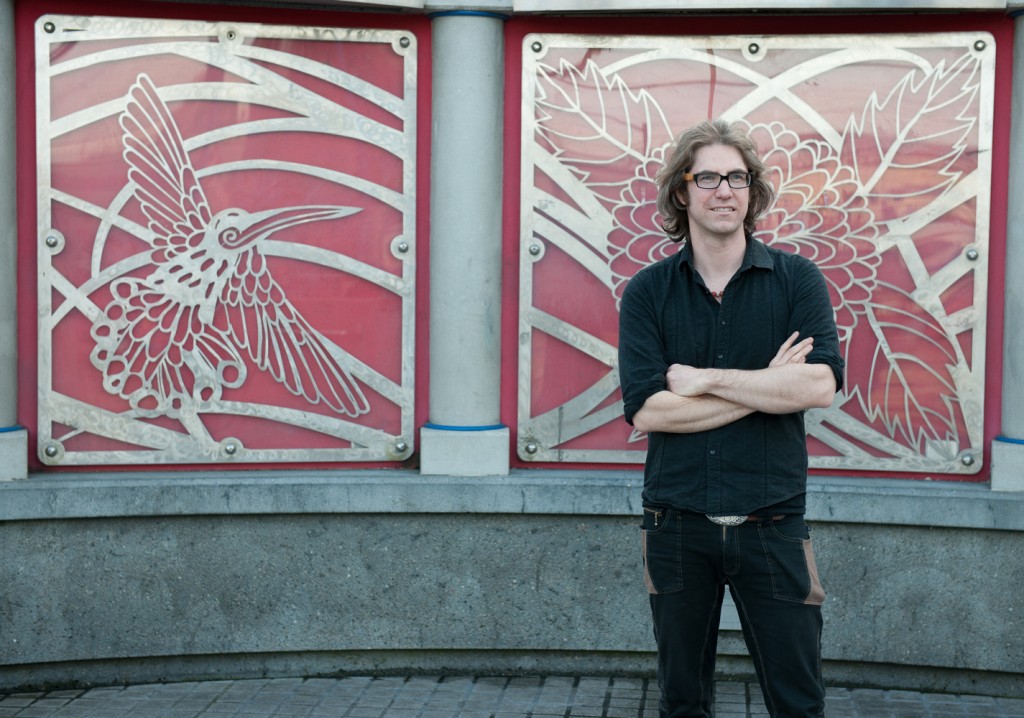 Man with long hair standing in front of a wall with steel cut artwork of leaves and a humming bird