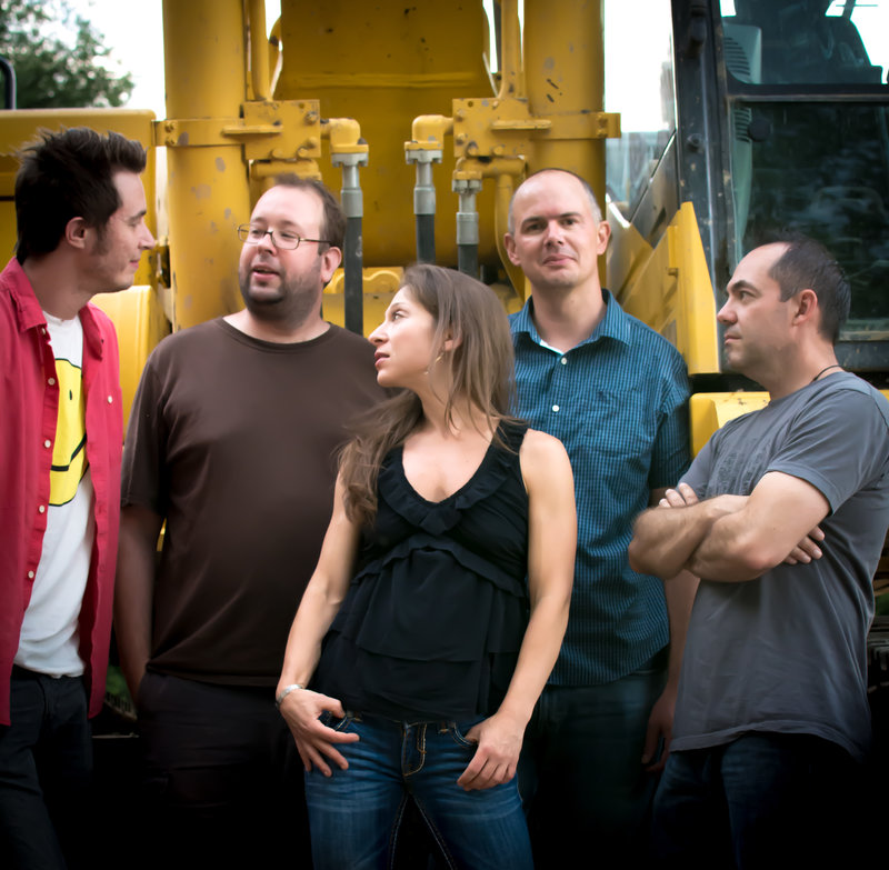 a band of five people stand in front of a yellow industrial looking background. all but one of the members is looking away from the camera.