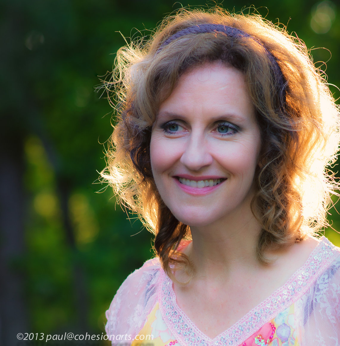 portrait of a woman to the right side of the image, she is smiling and gazing to her left, away from the viewer's gaze. sunlight hits her from beind, lighting her golden brown hair, falling beneath her chin but above her shoulders. she is wearing a multi-pastel colored top, and a purple braided headband.