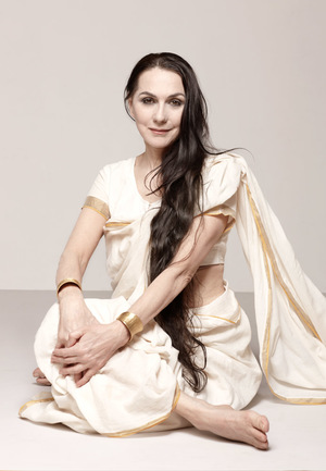 a yogini in cream sit smiling delicately at the viewer with her legs entwined, her hand across her knees, and her long brown hair reaching to her hips