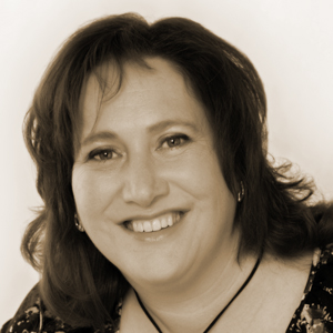 a dark haired, dark featured woman with a round face smiling and facing the camera directly. she is wearing a necklace, and has shoulder lengthed hair with a hint of bangs 