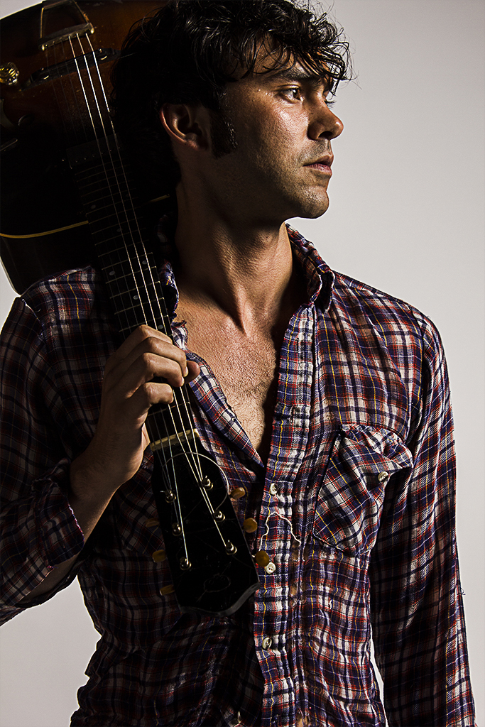 a dark featured man in a plaid shirt stands with a guitar slung over his right shoulder