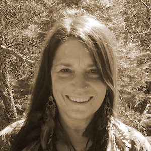 a thin faced brown haired woman smiles at the viewer in a backdrop of nature. she has a nose ring on her left nostril and large dangly earrings. her hair is long and she is wearing a collared, patterned top.