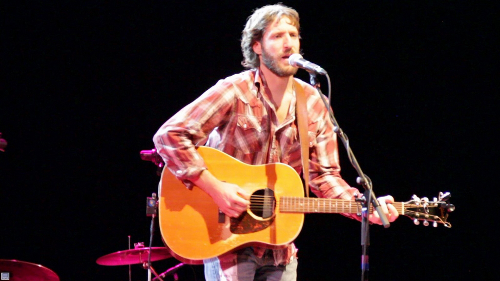 Todd Adelman stands on a stage playing a guitar, standing at a mic