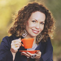 a woman with brown curcly hair holding a tea cup close to her face and grinning