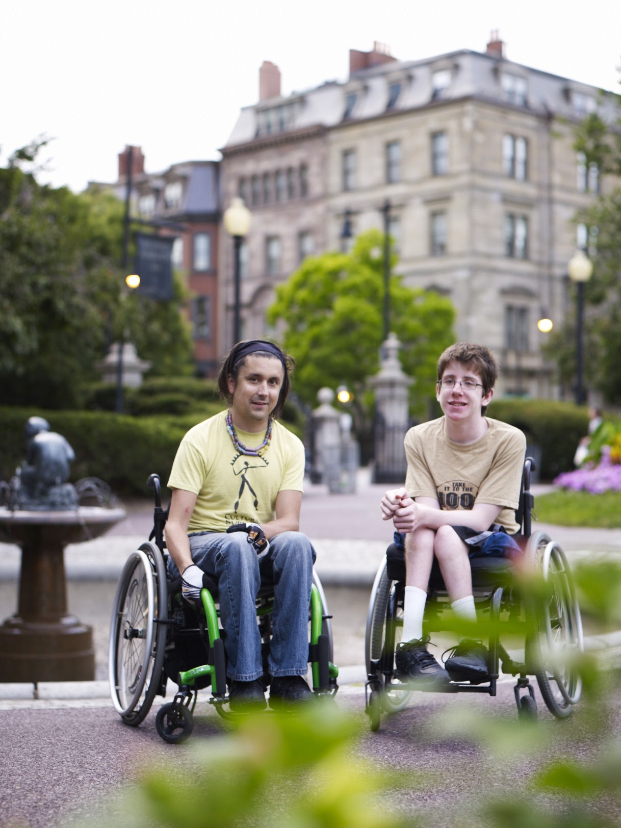 Tow people in wheel chairs smiling at camera, buildings in background
